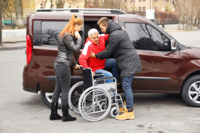 The driver and caregiver woman are helping handicapped senior man to sit in wheelchair